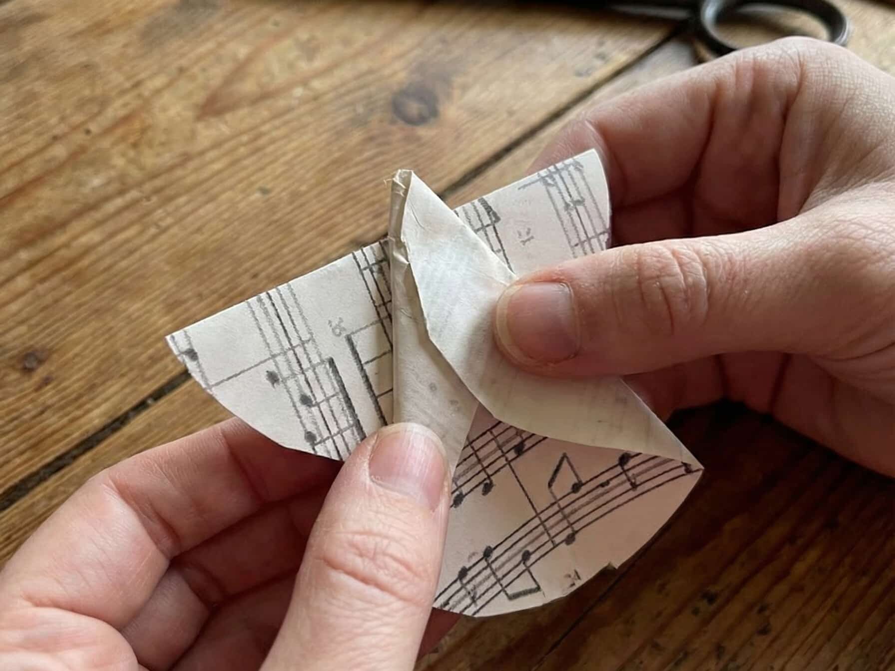 Výroba DIY dekorace anděla z vintage notového papíru na rustikálním dřevěném stole.