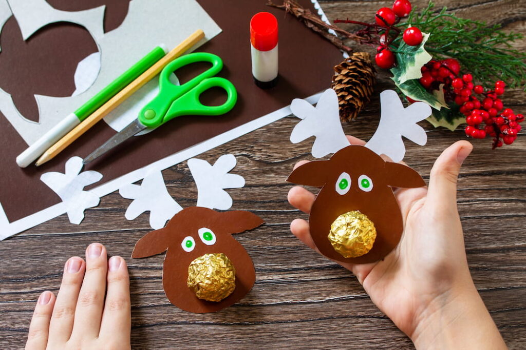 Dva dokončení papíroví sobíci leží na stole. Mají parohy, oči a místo nosu zlatou pralinku Ferrero Rocher.