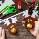 Dva dokončení papíroví sobíci leží na stole. Mají parohy, oči a místo nosu zlatou pralinku Ferrero Rocher.