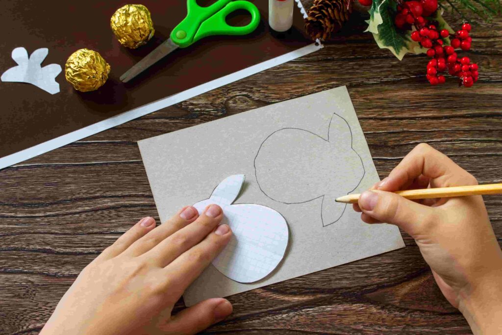 Dětské ruce obkreslují tužkou papírovou šablonu ve tvaru hlavy na tvrdý šedý karton.