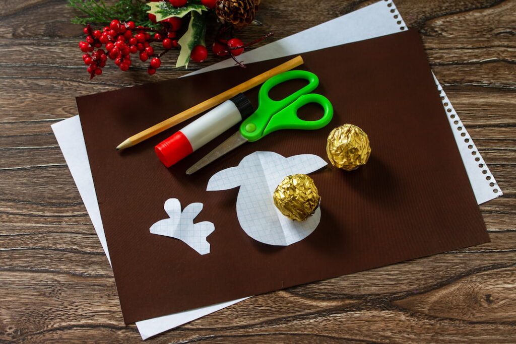 Na dřevěném stole leží hnědý papír, nůžky, tyčinkové lepidlo, tužka a dvě zlaté čokoládové pralinky.