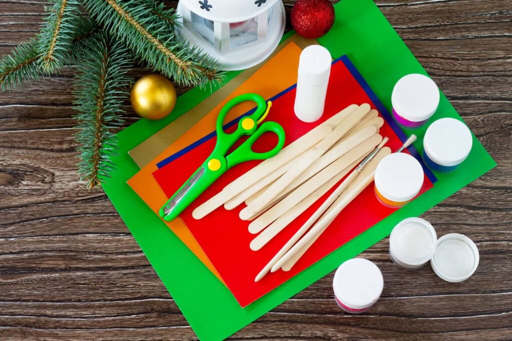 Detailní záběr na dřívka od nanuků, nůžky, lepidlo, štětce a barevné papíry na tmavém dřevěném pozadí s vánoční výzdobou.