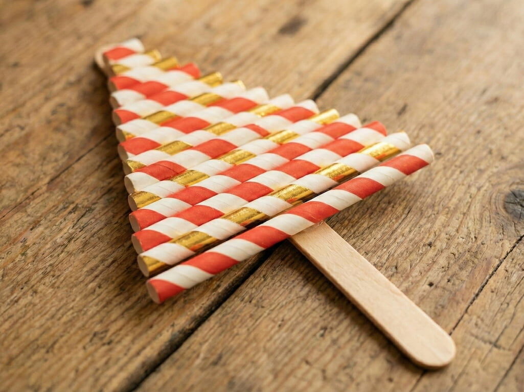 Hotový základ vánočního stromečku ležící na dřevěném stole. Stromeček je tvořen nalepenými papírovými brčky, kde se střídají červeno-bílé a zlato-bílé proužky.