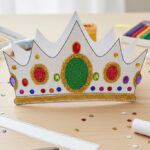 Hotová, barevně vyzdobená a vystřižená papírová koruna pro děti s nalepenými kamínky a třpytkami stojící na dřevěném stole.