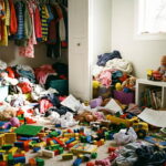 Neuklizený dětský pokoj plný barevného chaosu. Na světlém koberci v popředí jsou stovky rozházených kostek stavebnice, plastová auta, panenky a pokrčené papíry s kresbami. V pozadí je otevřená vestavěná skříň, ze které padají hromady dětského oblečení na podlahu.