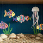 Dokončené papírové akvárium v krabici od bot s modrým pozadím, pískem na dně a barevnými třpytivými rybkami a medúzou zavěšenými na vlasci.
