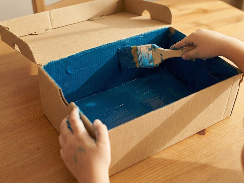 Dítě natírá vnitřek kartonové krabice modrou akrylovou barvou pomocí štětce, čímž vytváří pozadí pro akvárium.